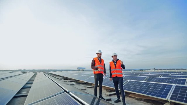 Solar generation technologies. Engineers standing on a flat roof with many solar panels. Workers in protective outfit and hard hats are talking on a modern solar farm.