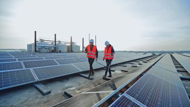New solar farm under the blue sky. Workers in special outfit walk and talk about sunny cells installation. Modern solar panels produce clean electricity.