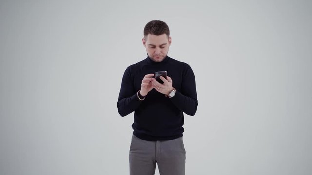 Handsome man with a cell phone isolated in studio. Portrait of a young man in black sweater and grey trousers looking in his phone.