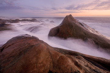 A beautiful misty seascape after sunset with clouds in the sky, with the waves splashing over the large rocks in the foreground, taken at Paternoster, South Africa.