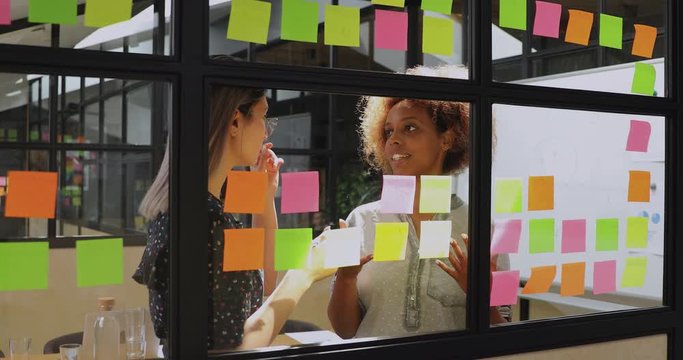 Smiling african american worker sharing project ideas with focused vietnamese colleague behind glass board. Motivated multiracial employees managing working processes on sticky paper notes in office.