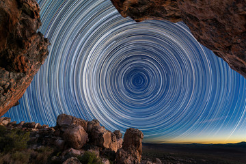 A beautiful night sky photograph with circular star trails framed by dramatic rocks in the...