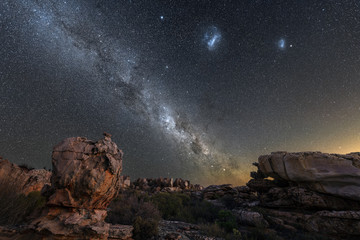 A beautiful night sky photograph showing the Milky Way and galactic centre, the Magellanic Clouds, with dramatic rocks in the foreground, taken in the Cederberg mountains in South Africa.