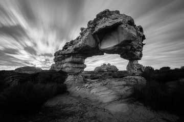 A dramatic black and white landscape photograph of an incredible rock arch before sunrise, with fast moving clouds against a moody sky, taken in the Cederberg mountains, South Africa.