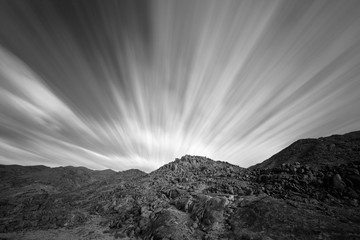 Un paysage de montagne aride en noir et blanc, utilisant une longue exposition pour brouiller les nuages en mouvement rapide avec une géologie fascinante et des formations rocheuses en arrière-plan, pris dans le Richtersveld en Afrique du Sud.