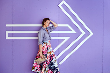 Young beautiful woman in a long skirt and a fashionable shirt with a pointing arrow on a delicate purple background.