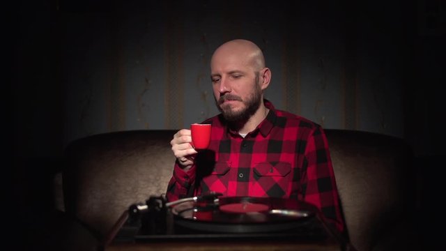Retro music expert bald caucasian white young hipster man with beard in red plaid shirt with red coffee cup enjoying retro turntable vintage vinyl player