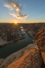 Obraz premium A beautiful vertical landscape view of the Augrabies Falls Gorge, mountains and river in South Africa, taken at sunset with a sunburst on the horizon and clouds in the blue sky.