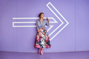 Young beautiful woman in a long skirt and a fashionable shirt with a pointing arrow on a delicate purple background.