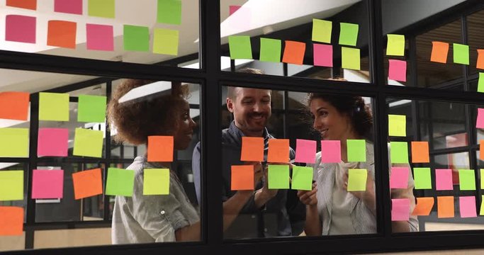 Excited happy young multiracial female colleagues sharing project ideas, joking, laughing with smiling male supervisor, standing behind window wall, writing notes on colorful paper stickers in office.
