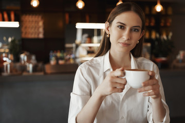 First coffee. Confident alluring young woman in white blouse, holding cup cappuccino, sitting in nice cafe with colleague after long day work, having lunch break, enjoying nice drink flavour