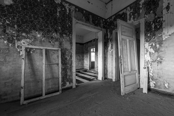 A black and white photograph inside an abandoned house with an open doorway, taken in the ghost town of Kolmanskop, Namibia.