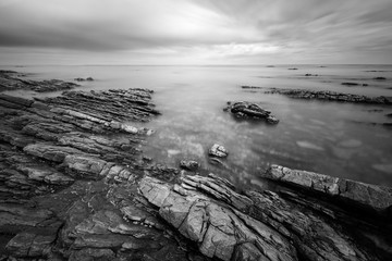 A black and white long exposure seascape of moving clouds and waves, creating a beautiful moody mist around the dramatic rocks in the foreground, taken in Port Nolloth, South Africa.