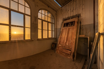 A dramatic photograph inside an abandoned house at sunrise, with a golden sunburst through broken windows and an ancient door buried in desert sand, taken in the ghost town of Kolmanskop, Namibia.