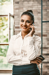 Fototapeta premium Smiling Business Woman Using Phone Looking At Camera In Office Interior, Portrait Of Beauty Women Speaking On Phone And Holding Another Phone In His Hand, Toned Image