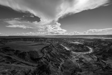 A black and white landscape taken at sunset on a stormy day on top of the arid and stark Fish River Canyon, Namibia, with the gorge and river in the foreground.