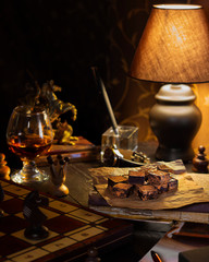 Moody brown composition of desk at man personal area at home with dark chocolate, glass of cognac, chess and lamp on it. Vertical wallpaper about relax time, hobby and food for gentlemen. Monochrome