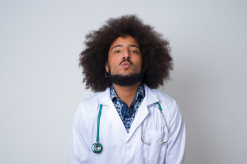 Pleasant looking young doctor male wearing medical uniform, keeps lips as going to kiss someone, has glad expression, grimace face. Standing indoors. Beauty concept.