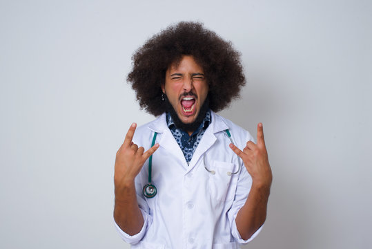 Born to rock this world. Joyful doctor man in medical uniform screaming out loud and showing with raised arms horns or rock gesture, expressing excitement of being on concert of band.