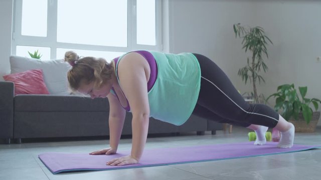 Plump woman barely doing push up exercise home, falling on floor, weak muscles