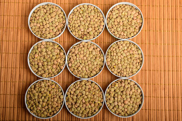 Top view of nine round bowls with many fresh organic green lentil beans on a wooden table in warm...