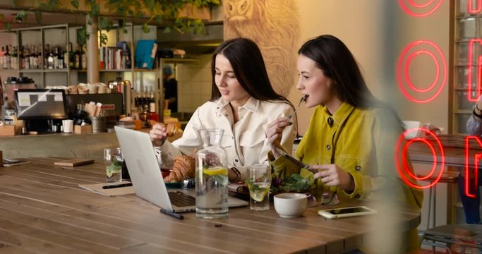 Pretty young women with long brown hair eating delicious food at local restaurant. Female friends in casual clothing enjoying morning breakfast. 