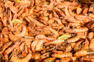 Close up of large portion of cooked shrimps with tomato sauce and yellow lemons in a large pan at a street food festival, ready to eat healthy seafood, beautiful orange monochrome outdoor background