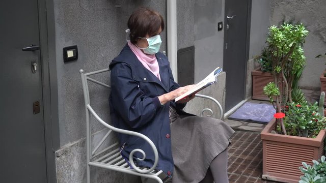 Europe - Italy - Milan - 70 year old lady with  mask on face to protect herself from the global epidemic of Novel Coronavirus n-cov 19 reads book during quarantine isolament