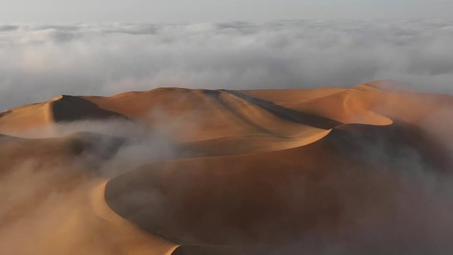 Aerial view from a drone flying over massive sand dunes that are surrounded by thick fog clouds at sunrise. Liwa desert, Abu Dhabi, United Arab Emirates.