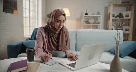 Beautiful young muslim girl in traditional hijab scarf is using her smartphone while working on project and positively smiling. Successful female freelancer or university student studying 4k footage - Powered by Adobe