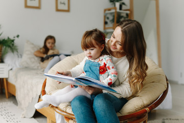 Portrait of a smiling young cute mother and two daughters reading a book relax in the sofa in a bright big white room