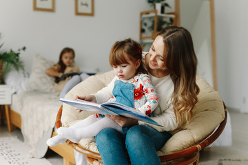Portrait of a smiling young cute mother and two daughters reading a book relax in the sofa in a...