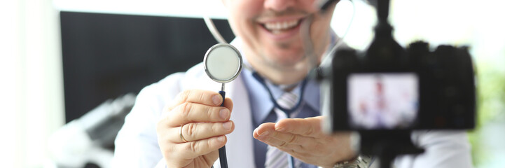 Obraz premium Portrait of smiling practitioner sitting in modern clinic office and showing important stethoscope vlog visitors. Cheerful man in white robe looking at camera. Clinic concept