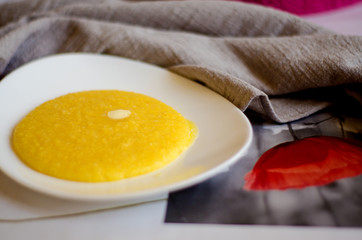 A portion of corn porridge with a piece of butter on a white plate next to a linen kitchen towel....