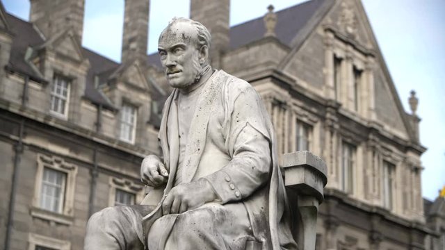 Statue of George Salmon in Trinity college, Dublin Ireland. Blue sky and giant building in background. Monument of influential Irish mathematician and Anglican theologian.