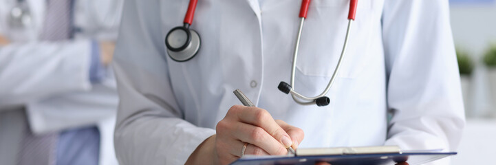 Close-up of female physician holding paper folder and writing something in documents. Doc wearing white medical uniform and stethoscope. Medicine and healthcare concept