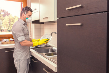 Man with the protective mask cleans the dishes at home.