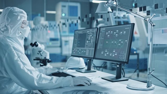 Electronics Factory Cleanroom: Engineer / Scientist in Coveralls Wors on Computer, Screen Shows Infographics and Software System Control UI, Developing Electronics for Medical Electronics
