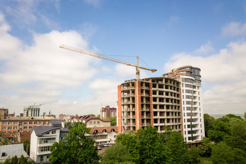 Fototapeta na wymiar Apartment or office tall building unfinished under construction among green tree tops. Tower cranes on bright blue sky copy space background.