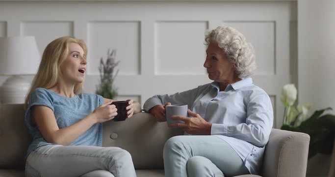 Happy young lady sitting on couch, chatting with pleasant middle aged 70s mother, drinking tea together. Smiling mature older woman holding cup of coffee, communicating with grown up daughter at home.