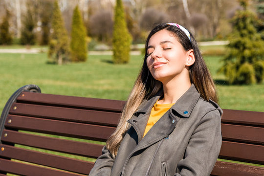 beautiful young hipster woman sitting on bench in park with eyes closed enjoying spring sun beams