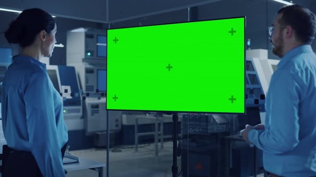 Office Meeting: Confident Female Engineer Talks to Project Manager, Watching Interactive Digital Whiteboard TV that Shows Green Screen Chroma Key Display. Modern Factory with Machinery
