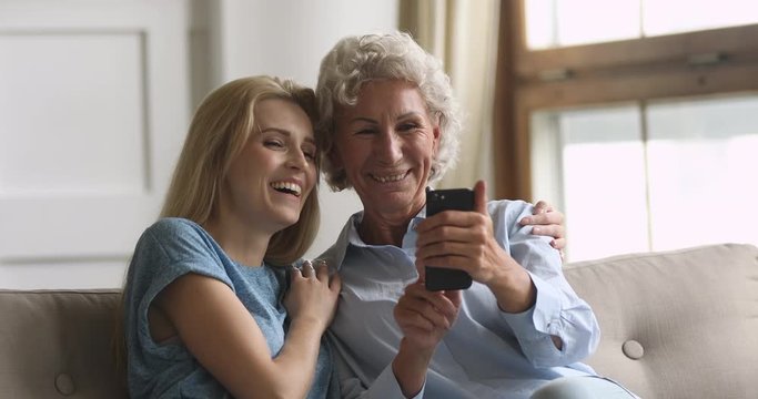 Happy elderly mature hoary woman trying to make selfie photo on smartphone with smiling blonde grown up daughter, sitting together on comfortable sofa. Overjoyed family having fun, using mobile apps.