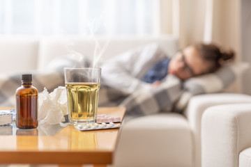 Young ill woman lying under blanket on a bed with seasonal illnes drinking hot tea with honey and lemon. She takes pills and cough sirup to push the disease