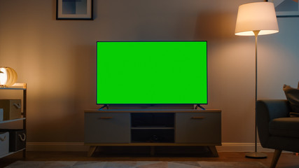 Shot of a TV with Horizontal Green Screen Mock Up. Cozy Evening Living Room with a Chair and Lamps...
