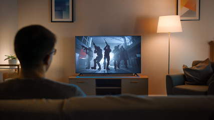 Young Man in Glasses is Sitting on a Sofa and Watching TV with an Action Movie with Soldiers. It's Evening and Room at Home Has Working Lamps. - obrazy, fototapety, plakaty