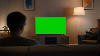 Young Man in Glasses is Sitting on a Sofa and Watching TV with Horizontal Green Screen Mock Up....