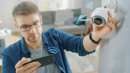 Young Man in Glasses Wearing a Blue Shirt is Adjusting a Modern Wi-Fi Surveillance Camera with Two Antennas on a White Wall at Home. He's Checking the Video Feed on his Smartphone. - obrazy, fototapety, plakaty