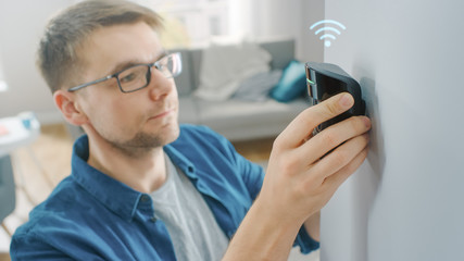 Young Man in Glasses Wearing a Blue Shirt is Hanging a Modern Movement Detector Unit with Digital Icon of Wireless Connection Above it on a White Wall at Home. Device Blinks with Green Led Light.