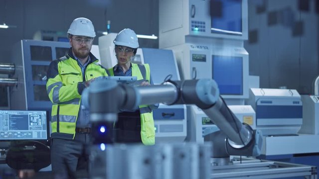 Factory Workshop: Professional Female Engineer, Male Machinery Operator Use Industrial Digital Tablet Computer to Work and Program Robot Arm for Production Line. High-Tech Facility with CNC Machines